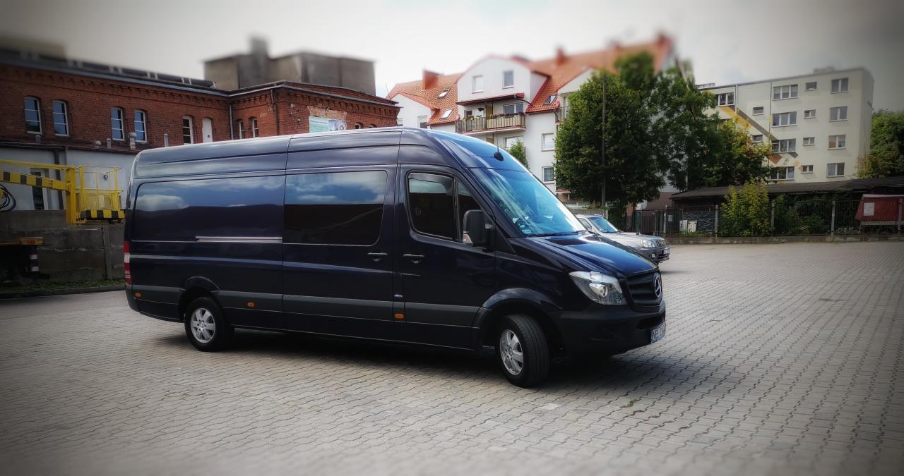 Czarny bus Mercedes-Benz Sprinter na parkingu z kostki, budynki w tle. Pojazd o podwyższonym dachu, z długim rozstawem osi, idealny do transportu osób lub towarów.
