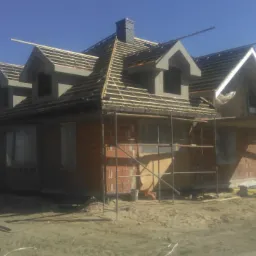 Dom jednorodzinny w trakcie budowy: widoczne murowane ściany z czerwonej cegły, konstrukcja dachu z drewnianych łat, szare lukarny, rusztowania wokół budynku, teren budowy z piaskiem i gruzem.