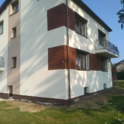 Dwukondygnacyjny dom z balkonami i brązowymi panelami dekoracyjnymi na elewacji, widok z zewnątrz w słoneczny dzień.