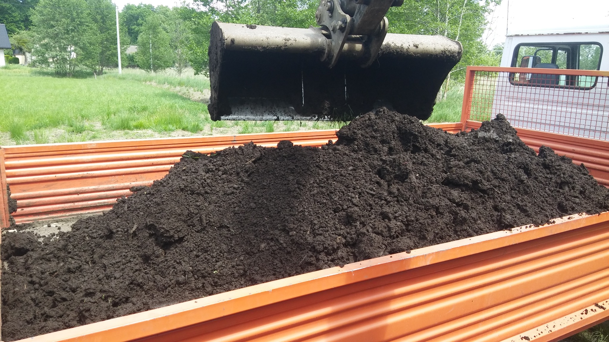 Łyżka koparki wysypuje ciemną ziemię do pomarańczowej skrzyni ciężarówki, widoczne tło zieleni i budynki.