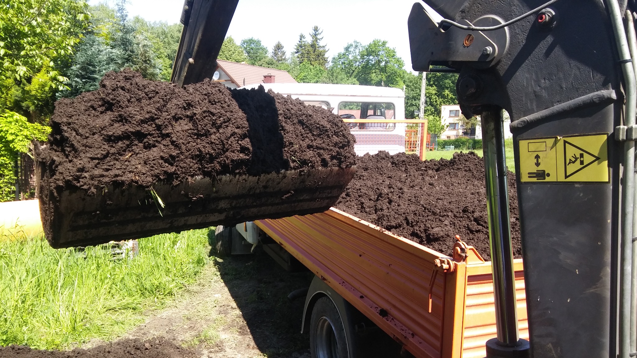 Łyżka koparki z ciemną ziemią wysypuje urobek na pomarańczową skrzynię samochodu ciężarowego, w tle trawa i zabudowania.