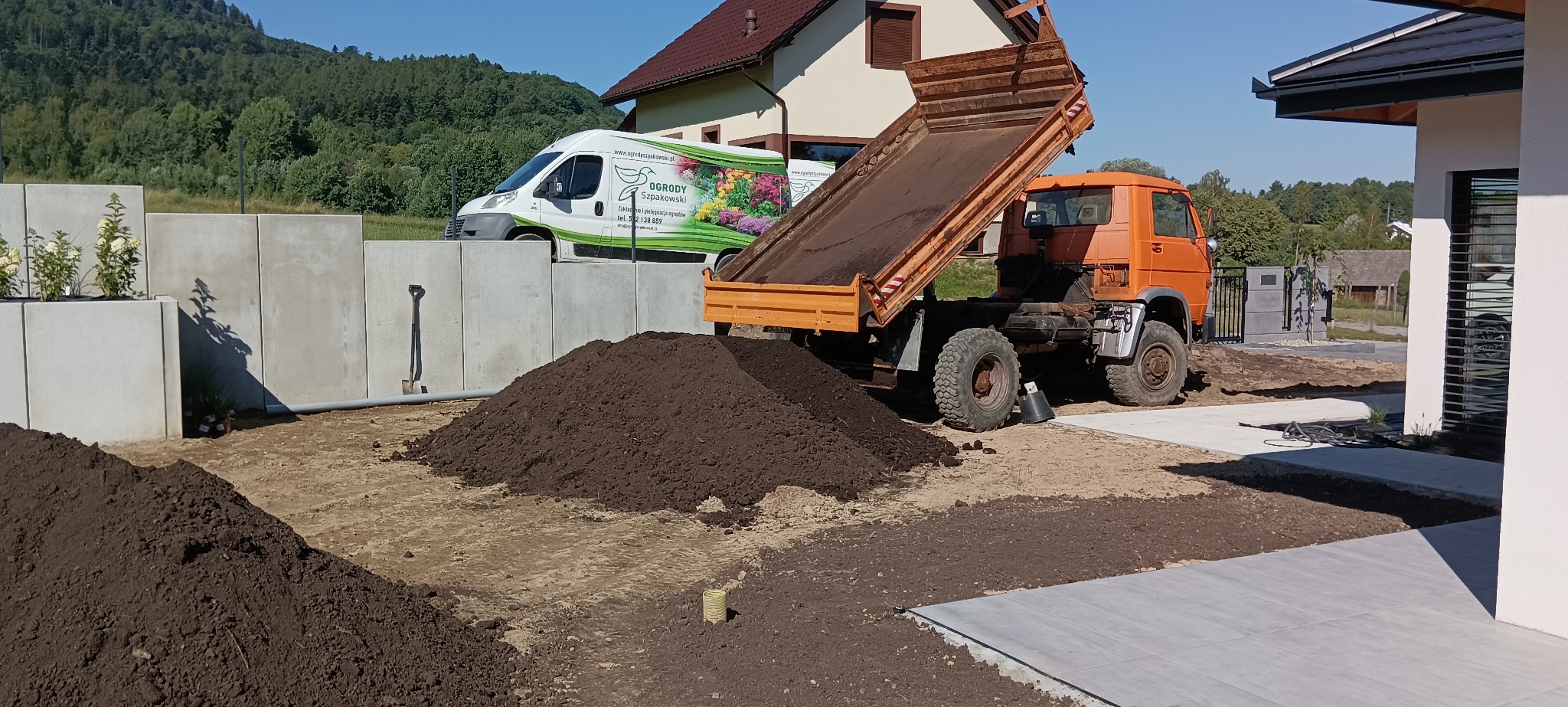 Pomarańczowa wywrotka z podniesioną skrzynią stoi na działce obok domu, z tyłu widoczny bus z reklamą firmy ogrodniczej i betonowe ściany z roślinami.