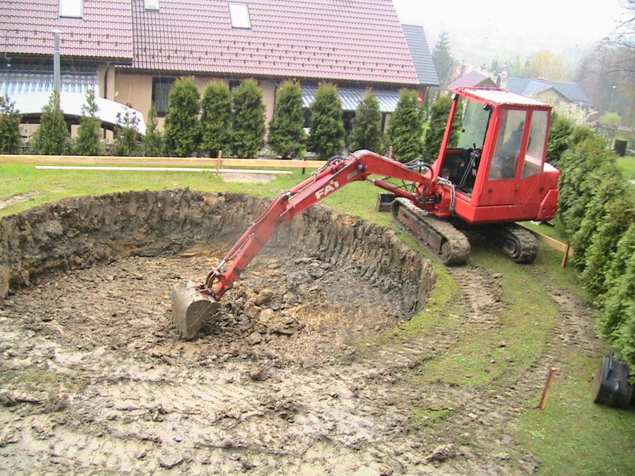 Czerwona mini koparka na gąsienicach w trakcie pogłębiania okrągłego wykopu w ziemi, prawdopodobnie pod staw, z widocznymi śladami opon i zabudowaniami w tle.