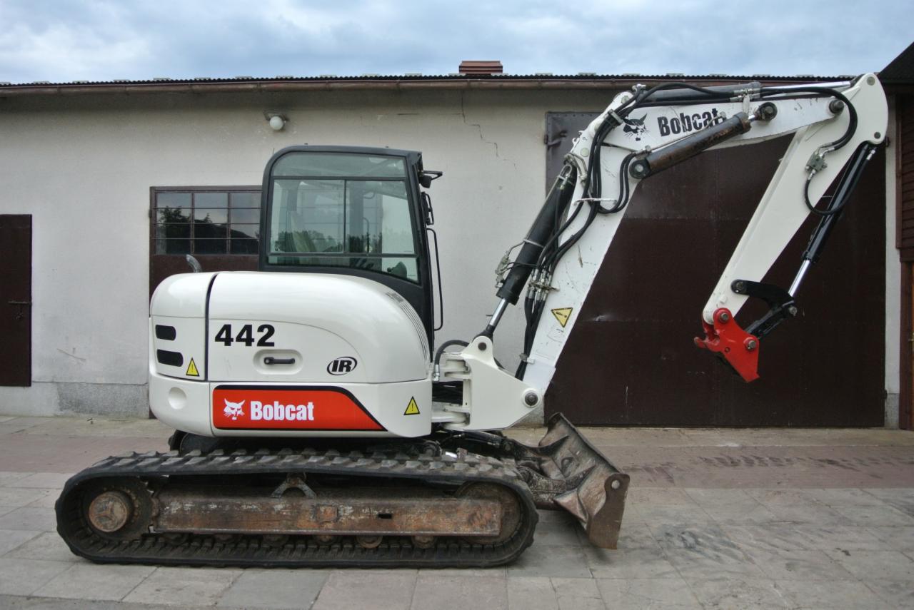 Biała mini koparka Bobcat 442 na gumowych gąsienicach stoi na szarym bruku przed budynkiem z brązowymi wrotami garażowymi.