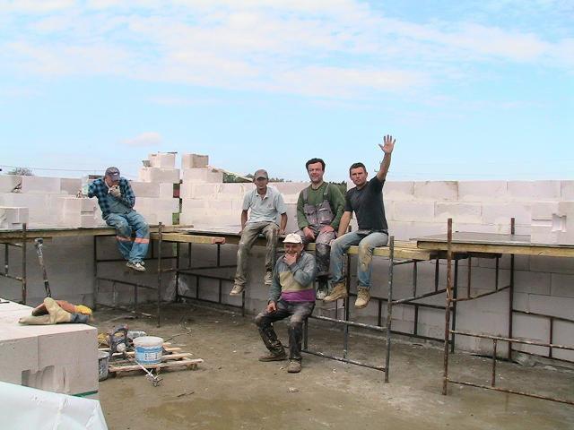 Grupa pięciu robotników w ubraniach roboczych odpoczywa na rusztowaniu podczas budowy fundamentów z białych bloczków, jeden z nich macha ręką.