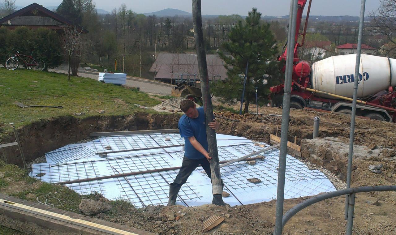 Pracownik budowlany w gumowych butach trzyma wąż pompy do betonu nad wykopem z ułożoną siatką zbrojeniową na białej izolacji, w tle betoniarka i krajobraz wiejski.