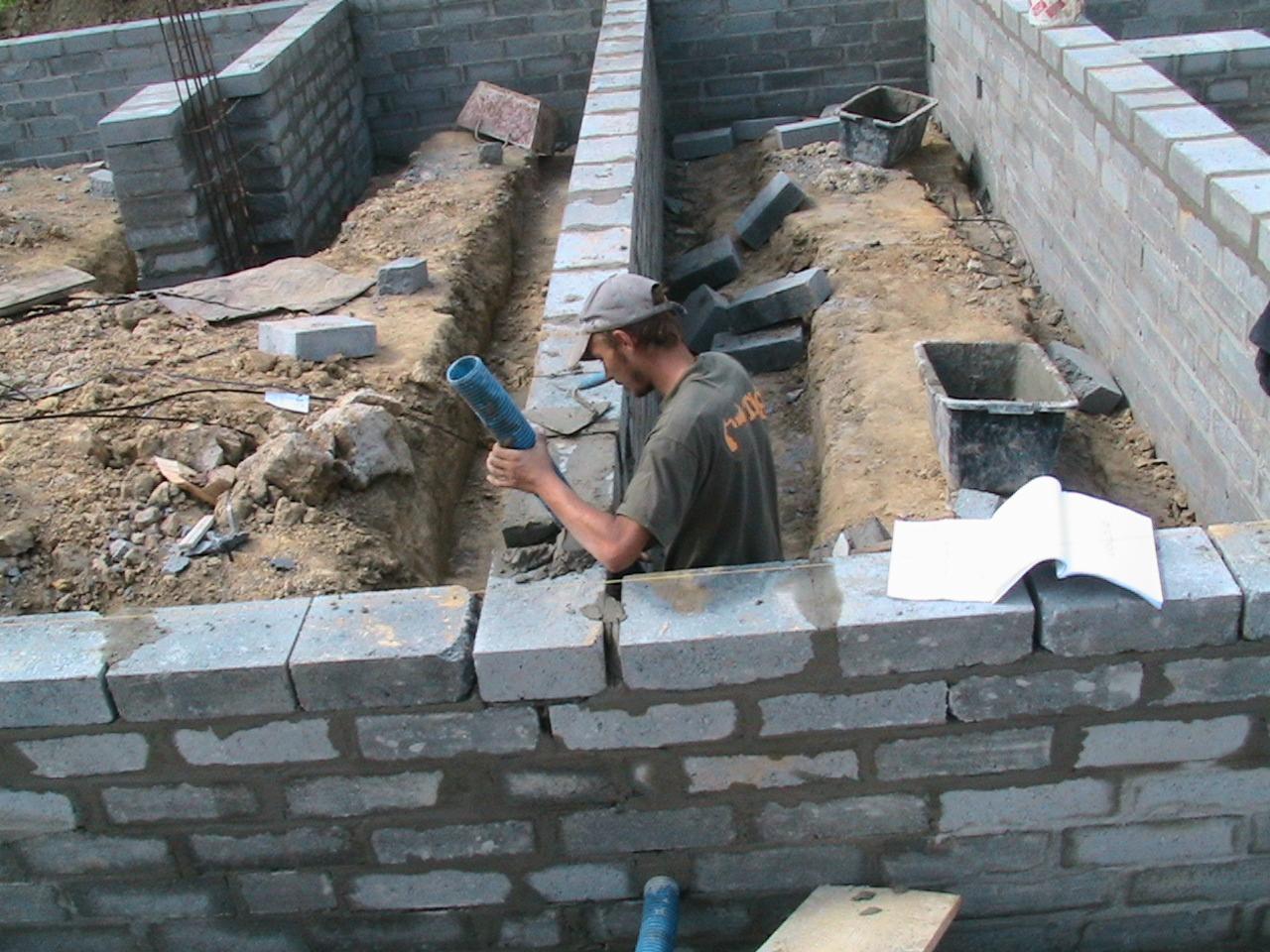 Budowa fundamentów: murarz układa bloczki betonowe, trzymając niebieską rurę w wykopie, widoczne szalunki i plan budowy.