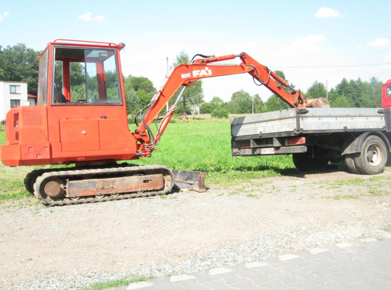 Czerwona koparka gąsienicowa FAI o wadze 3,7 tony w Skoczowie, z operatorem, załadowuje urobek na skrzynię samochodu ciężarowego. Widoczne ślady użytkowania na maszynie.