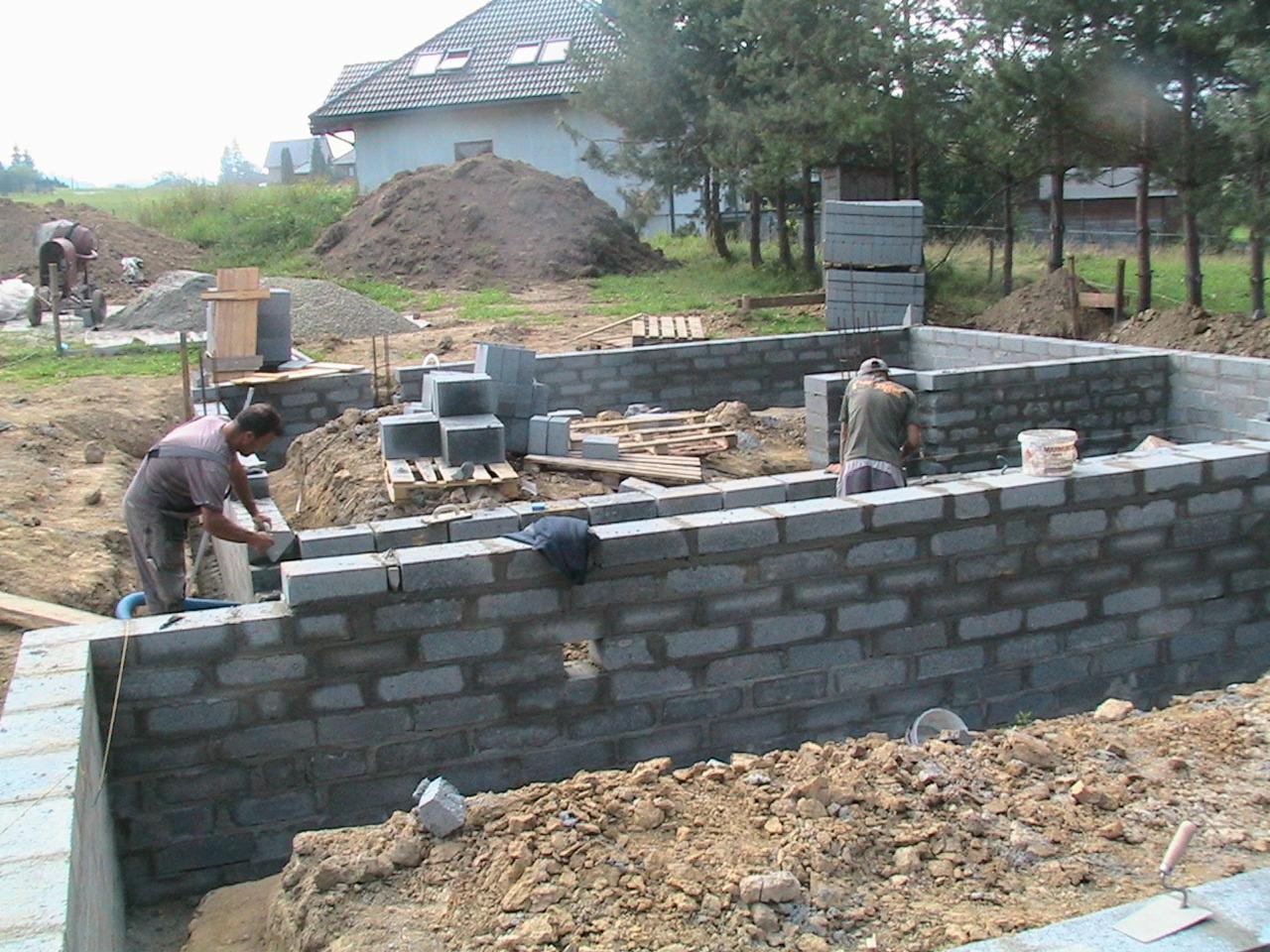 Budowa fundamentów domu z bloczków betonowych: murarz układa kolejne warstwy, widoczny wykop, zapasy bloczków na palecie, mieszalnik do betonu w tle.