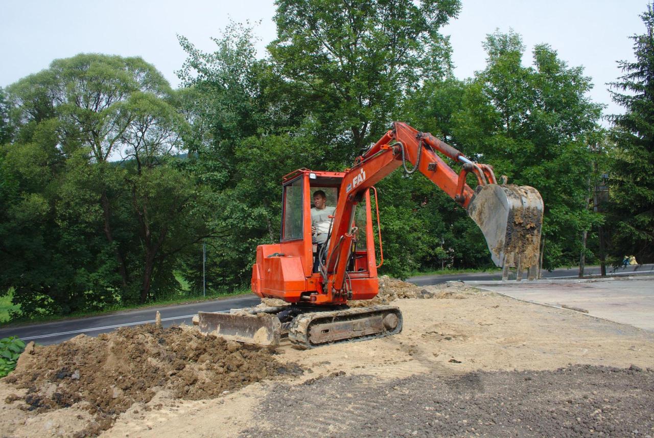 Mała, pomarańczowa koparka gąsienicowa FAI, z operatorem w kabinie, nabiera ziemię łyżką w pobliżu asfaltowej drogi i zielonych drzew.