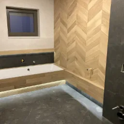 Nowoczesna łazienka w trakcie wykańczania: jasne drewnopodobne płytki ułożone w jodełkę na ścianie, szara podłoga, wanna obudowana drewnem z podświetleniem LED, okno w białej ścianie z szarymi...