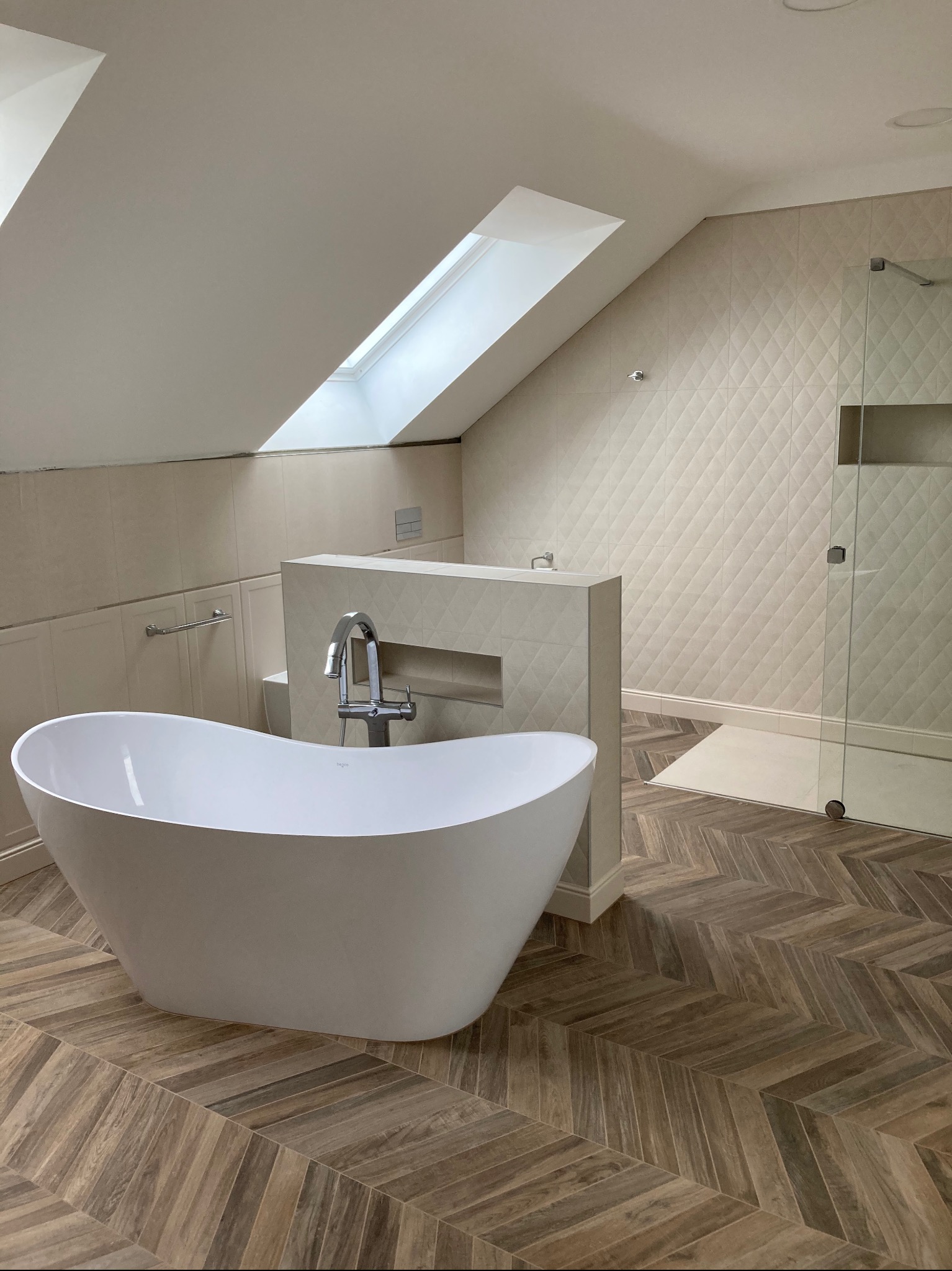 Nowoczesna łazienka na poddaszu z wolnostojącą białą wanną, mozaikową ścianą pod skosem i kabiną prysznicową z odpływem liniowym, drewnopodobna podłoga ułożona w jodełkę.
