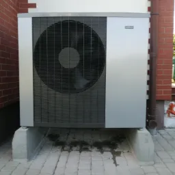 Zewnętrzna jednostka pompy ciepła zamontowana na betonowych podstawach przy ścianie budynku z cegły i tynkiem, widoczny brązowy spust wody deszczowej.