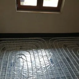 Białe rury ogrzewania podłogowego ułożone na folii z nadrukowaną siatką, widok z góry, czarna folia zabezpieczająca przy ścianie, małe okno w tle.