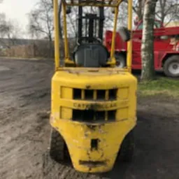 Żółty, używany wózek widłowy marki Caterpillar, sfotografowany na zewnątrz na nierównym, ziemistym podłożu. Widoczne ślady użytkowania i zabrudzenia na karoserii. W tle fragment czerwonego pojazdu...