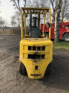 Żółty, używany wózek widłowy marki Caterpillar, sfotografowany na zewnątrz na nierównym, ziemistym podłożu. Widoczne ślady użytkowania i zabrudzenia na karoserii. W tle fragment czerwonego pojazdu...