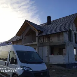 Nowo powstający dom jednorodzinny z widoczną konstrukcją dachu i częściowo ułożonym pokryciem z dachówki ceramicznej, na pierwszym planie biały bus firmy budowlanej.