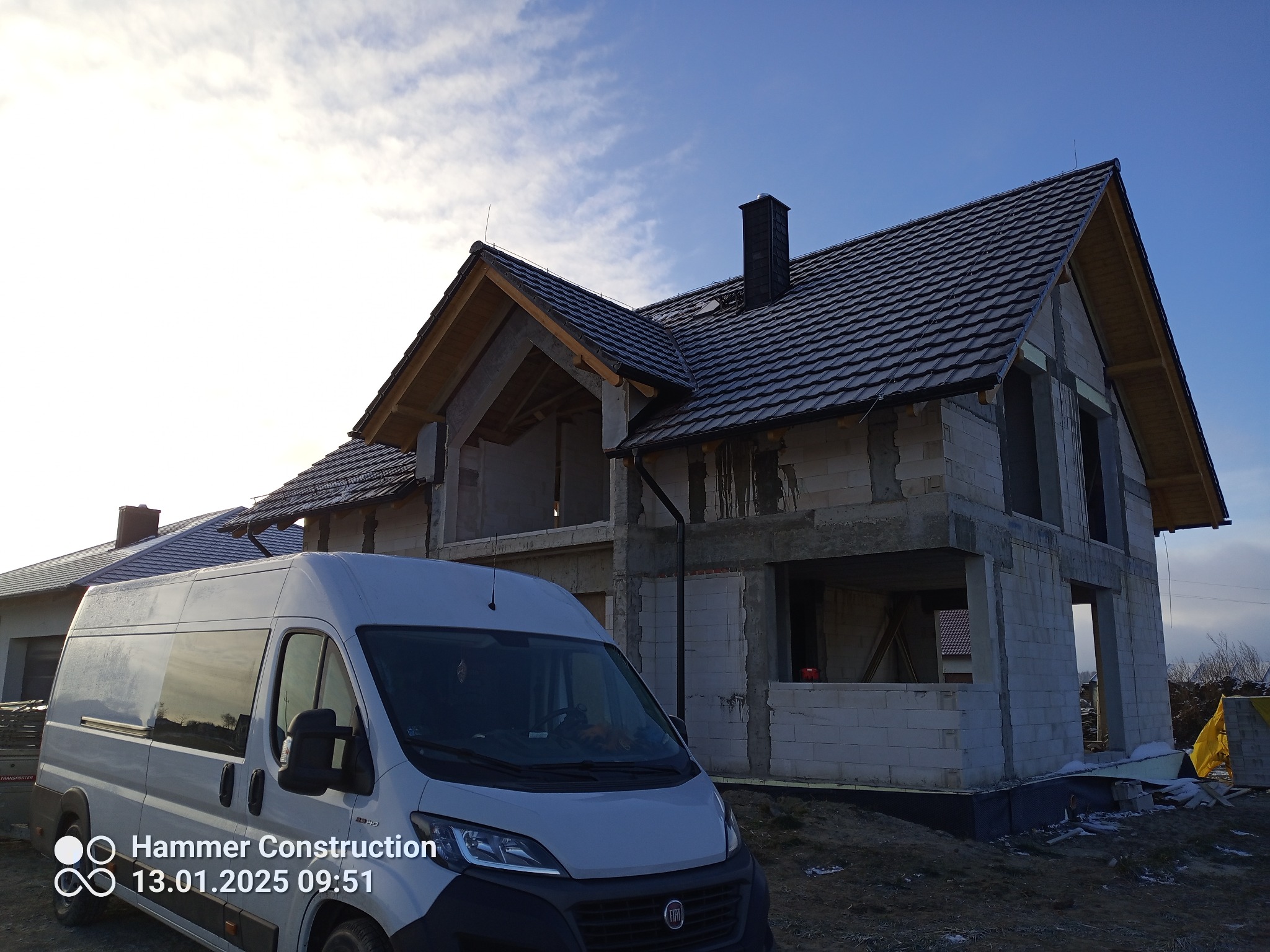 Nowo powstający dom jednorodzinny z widoczną konstrukcją dachu i częściowo ułożonym pokryciem z dachówki ceramicznej, na pierwszym planie biały bus firmy budowlanej.