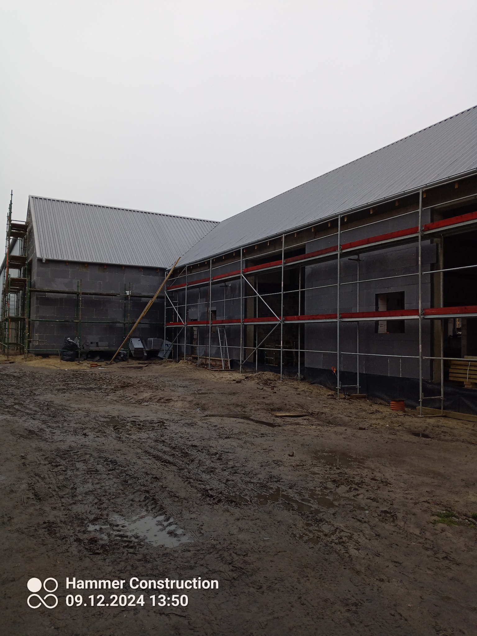 Budynek w trakcie budowy z szarą blachą na dachu, otoczony rusztowaniami i błotnistym terenem.