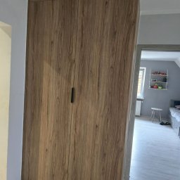 AR-GRA WYKOŃCZENIA WNĘTRZ ARKADIUSZ GRABOWSKI - Wysoka szafa z drewnopodobnymi frontami wkomponowana we wnękę ściany, widok na pokój dziecięcy z łóżkiem i półką na ścianie.