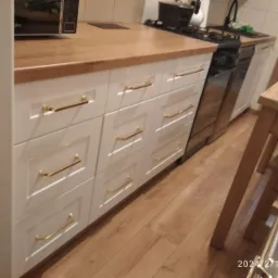 Białe szafki kuchenne z drewnianym blatem i złotymi uchwytami, widok na podłogę z paneli i fragment kuchenki gazowej.