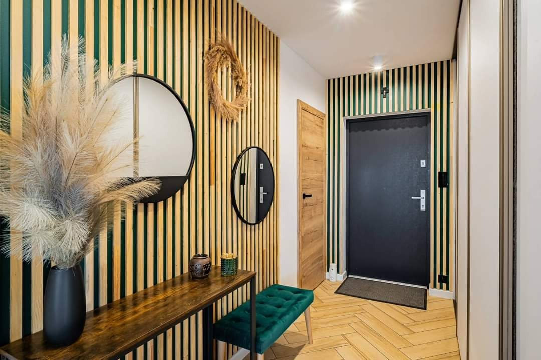 Nowoczesny przedpokój z panelami ściennymi w odcieniach zieleni i beżu, okrągłe lustra, drewniany stolik z dekoracjami i zielonym pufem, parkiet ułożony w jodełkę.