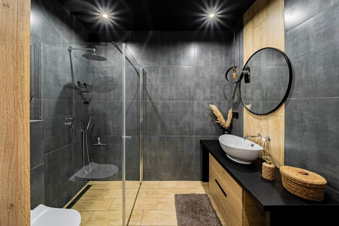 Nowoczesna łazienka z kabiną prysznicową typu walk-in, szarą glazurą, drewnianymi akcentami i okrągłym lustrem podwieszonym na tle drewnianej ściany.