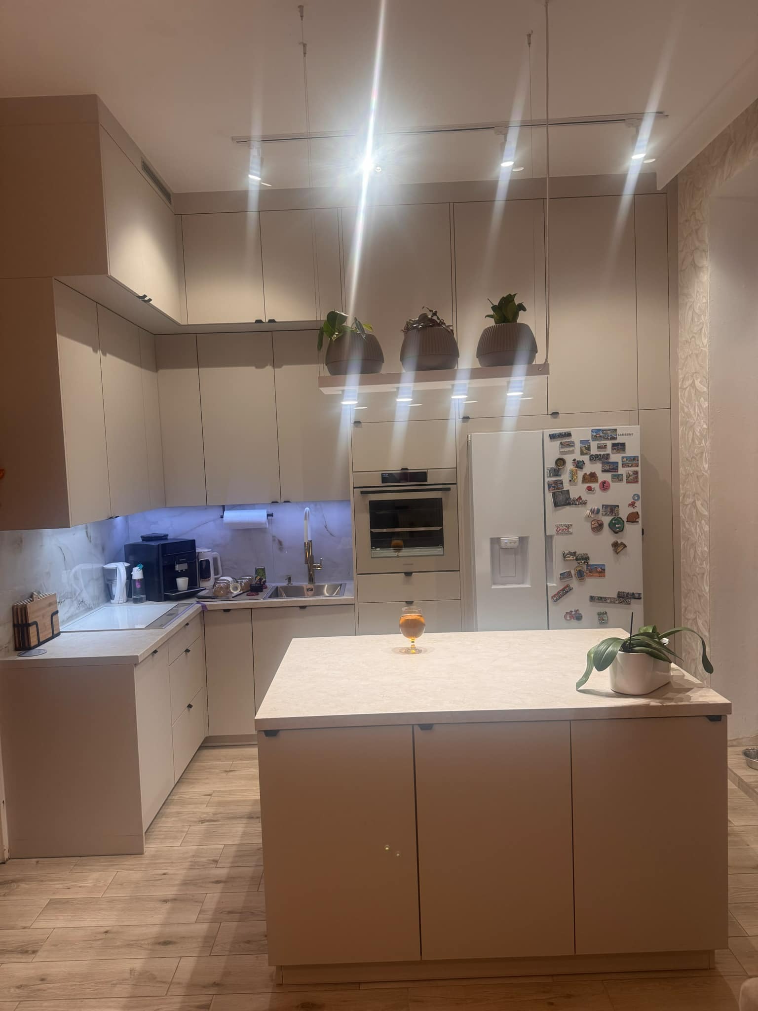 Jasna kuchnia z wyspą, oświetleniem punktowym i marmurowym backsplashiem, dekorowana roślinami w doniczkach i magnesami na lodówce.