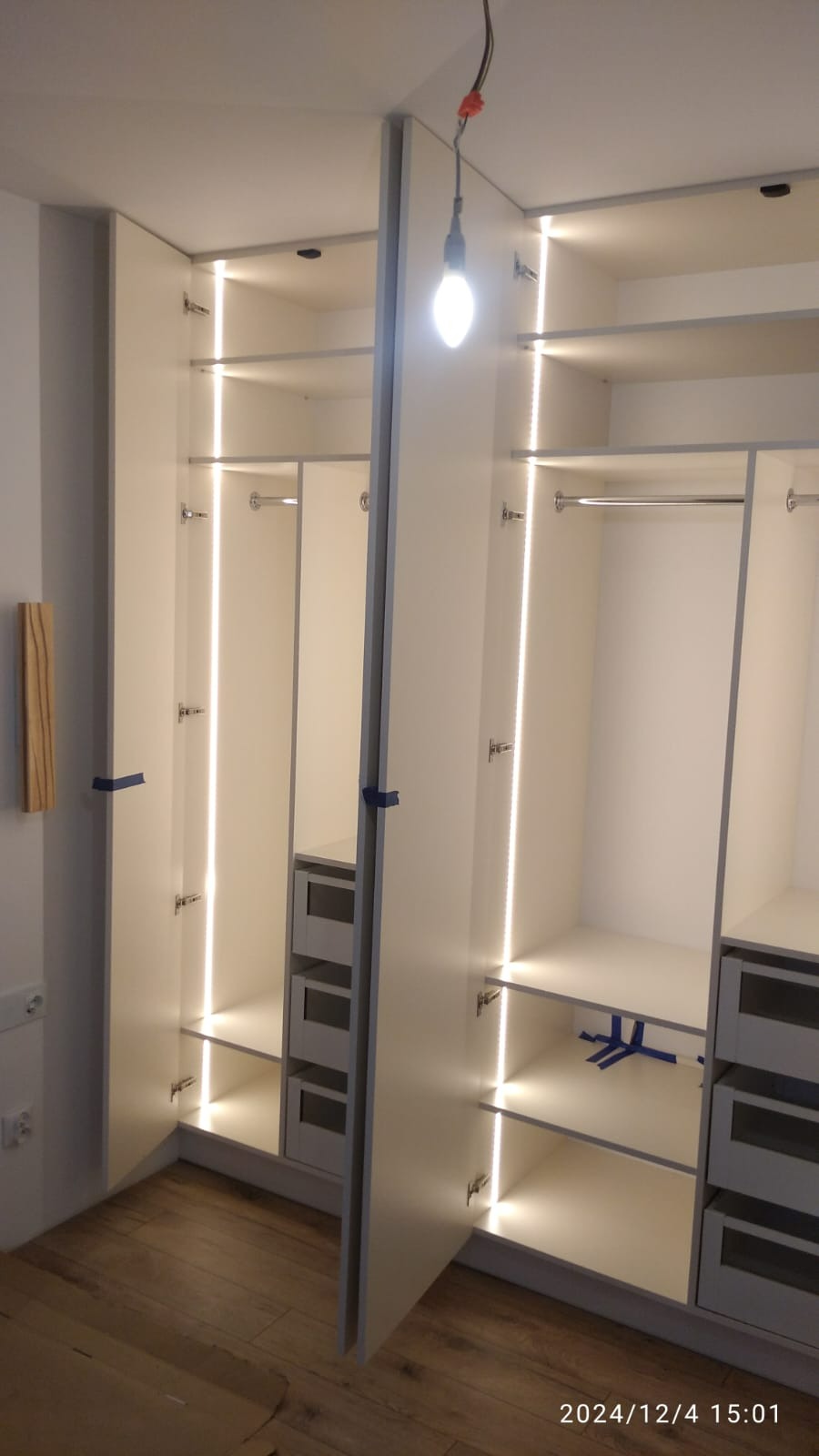 Wnętrze szafy wnękowej z oświetleniem LED, widoczne półki, drążek na ubrania, szuflady i drewniana podłoga. Drzwi szafy częściowo otwarte, zabezpieczone niebieską taśmą malarską.