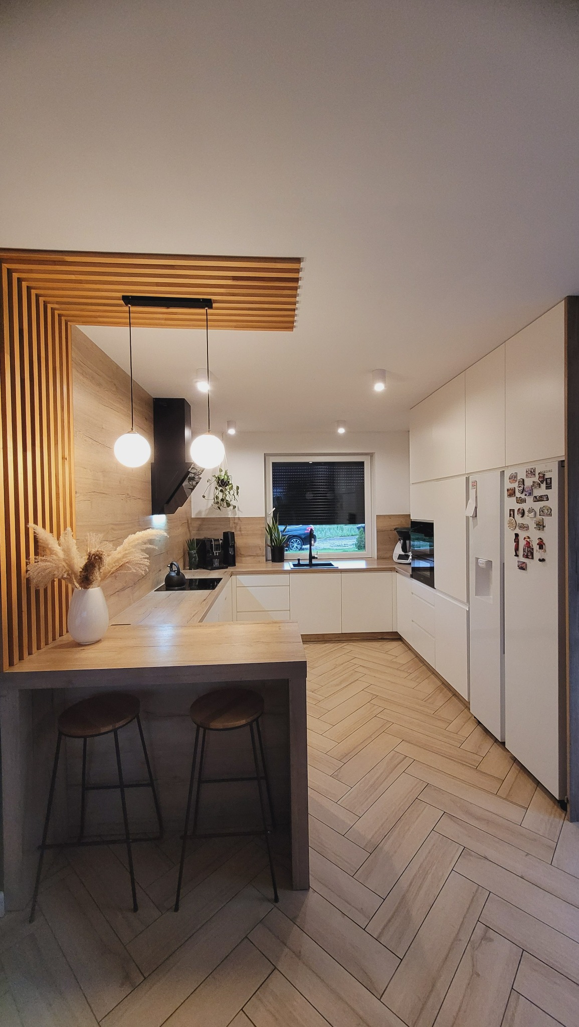 Nowoczesna kuchnia z drewnianymi akcentami: lamele na ścianie i suficie, białe szafki, wyspa z hokerami i podłoga ułożona w jodełkę.