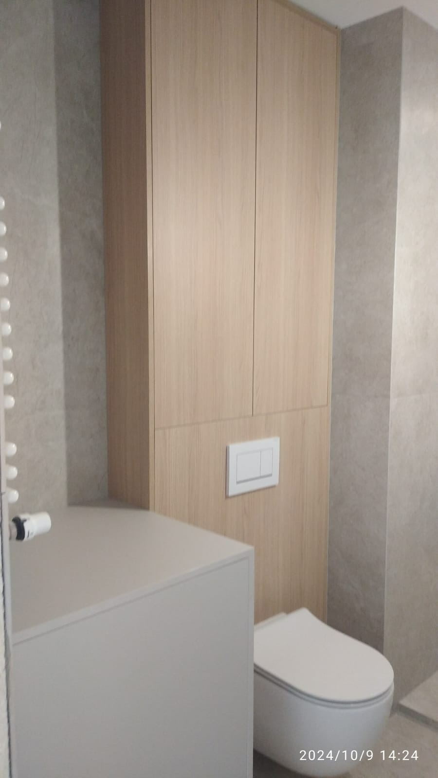 Nowoczesna łazienka z podwieszaną toaletą, zabudową meblową z drewnopodobnym fornirem i szarą płytką na ścianach.