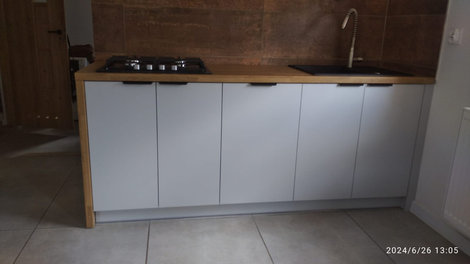 Szare szafki kuchenne z drewnianym blatem, na którym umieszczono płytę grzewczą i zlew z baterią. Tło stanowi brązowa ściana imitująca kamień i fragment otwartego pokoju.