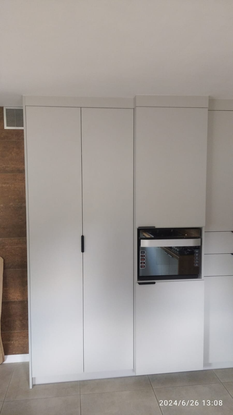 Biała zabudowa kuchenna z wbudowanym piekarnikiem i szafkami, minimalistyczny design, widoczna brązowa ściana w tle.