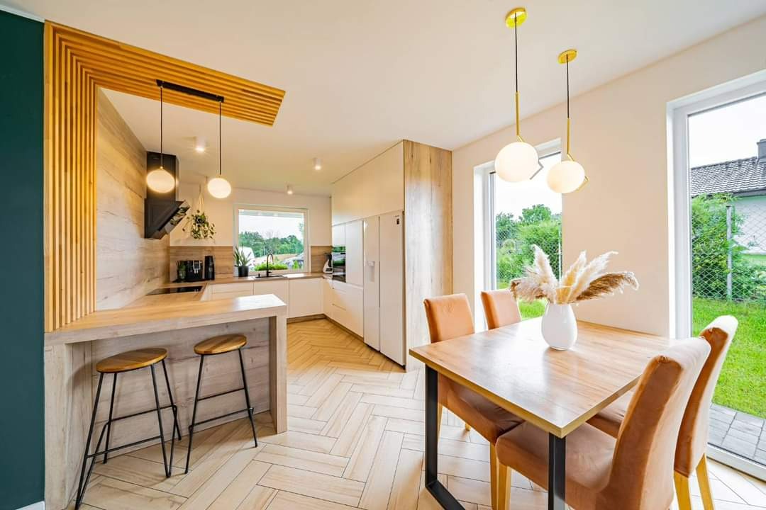 Jasna kuchnia z drewnianymi akcentami, parkiet ułożony w jodełkę, wyspa z hokerami i stół jadalny z krzesłami, oświetlona wiszącymi lampami z okrągłymi kloszami.