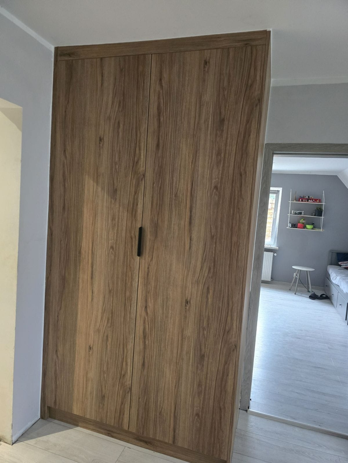 Wysoka szafa z drewnopodobnymi frontami wkomponowana we wnękę ściany, widok na pokój dziecięcy z łóżkiem i półką na ścianie.