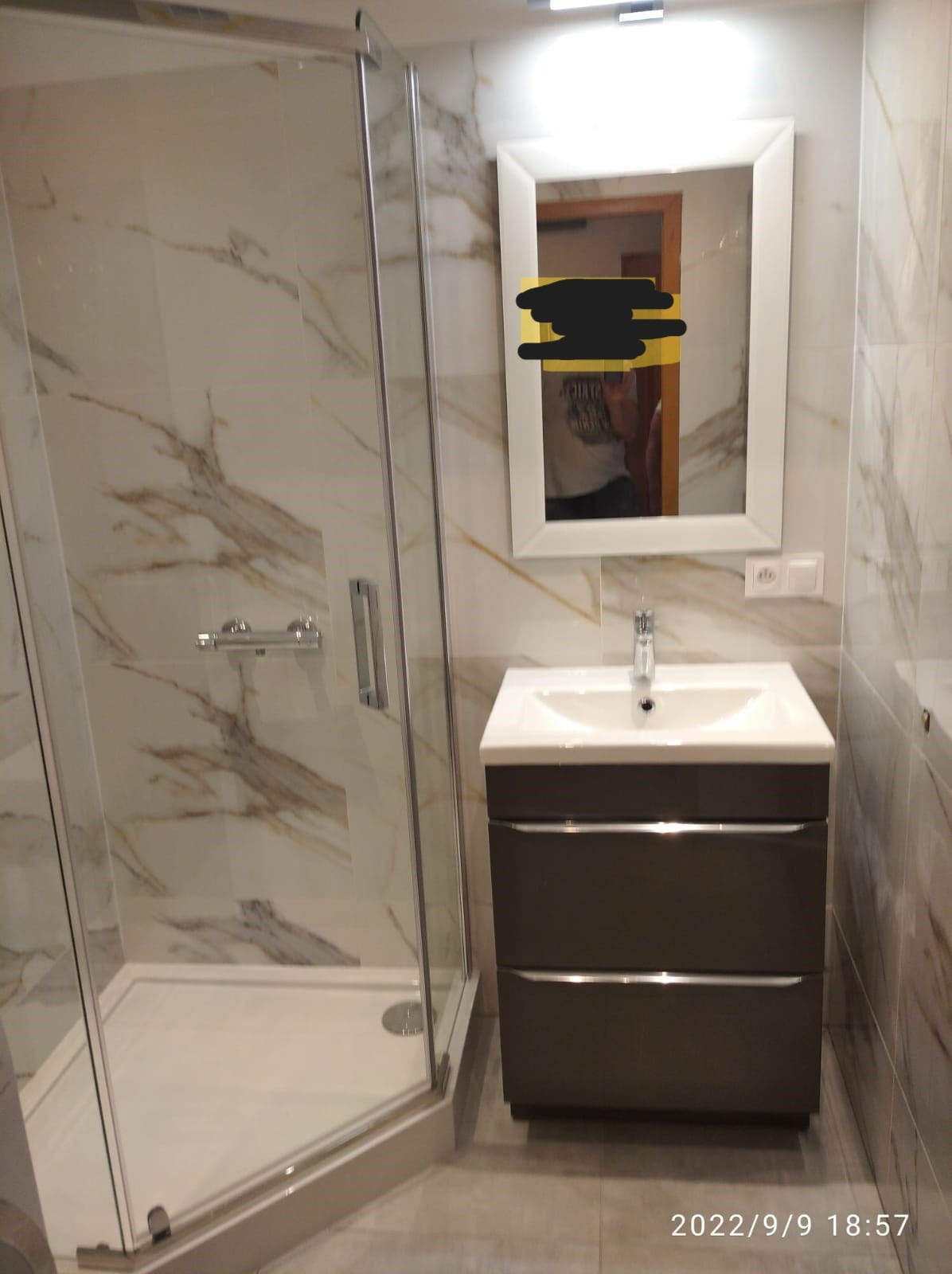 Nowoczesna łazienka z kabiną prysznicową typu walk-in, umywalką z szafką w kolorze grafitowym i lustrem z oświetleniem LED, wykończona płytkami imitującymi marmur.