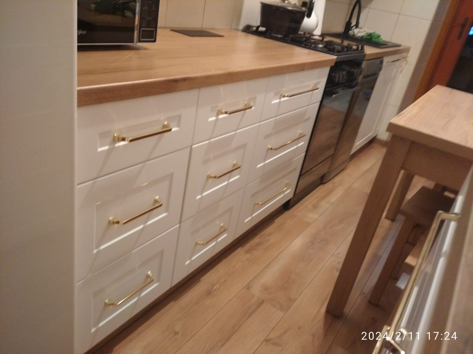 Białe szafki kuchenne z drewnianym blatem i złotymi uchwytami, widok na podłogę z paneli i fragment kuchenki gazowej.