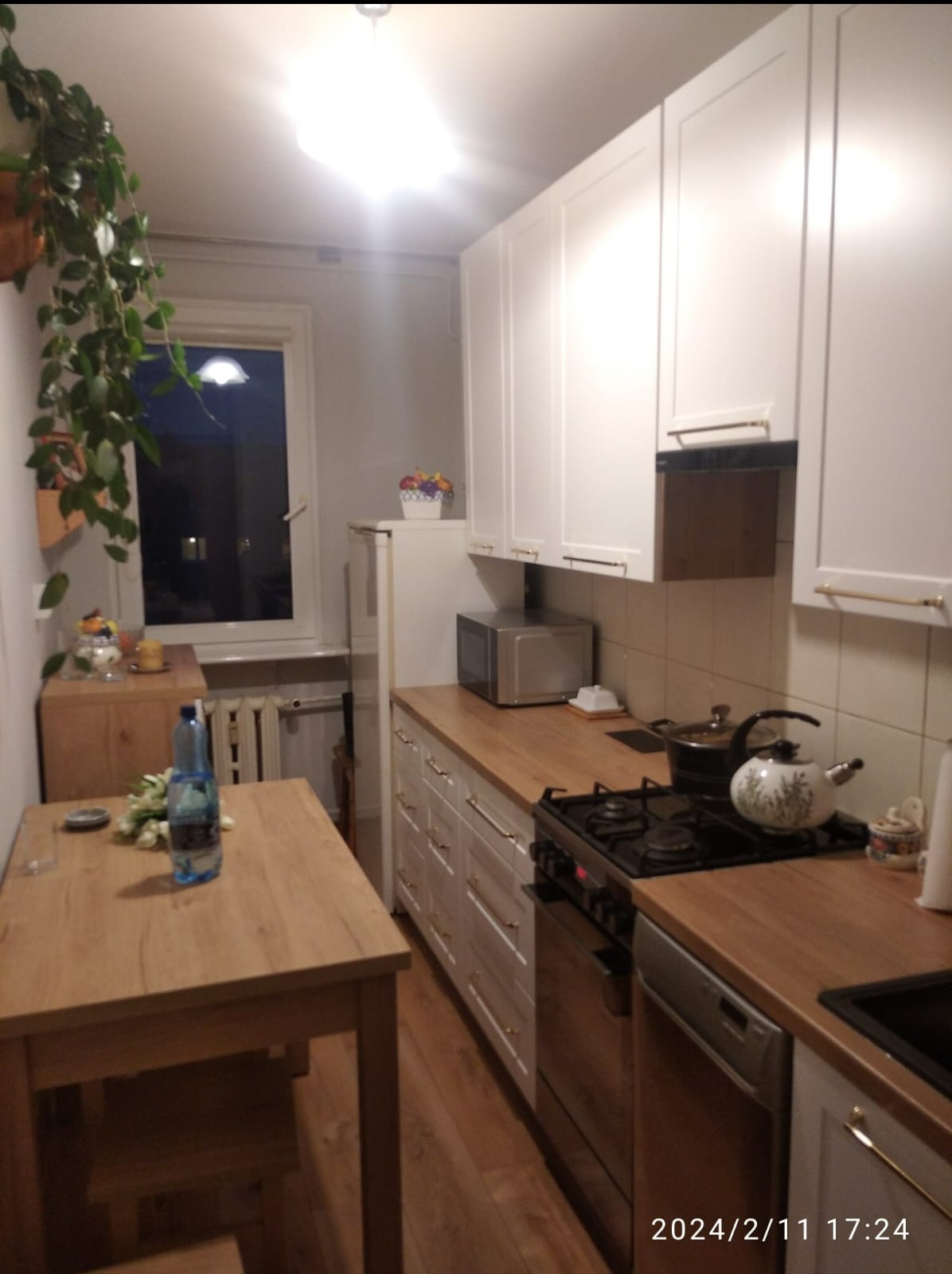 Jasna kuchnia z drewnianym blatem, białymi szafkami i stołem z butelką wody, widok na okno z rośliną doniczkową.