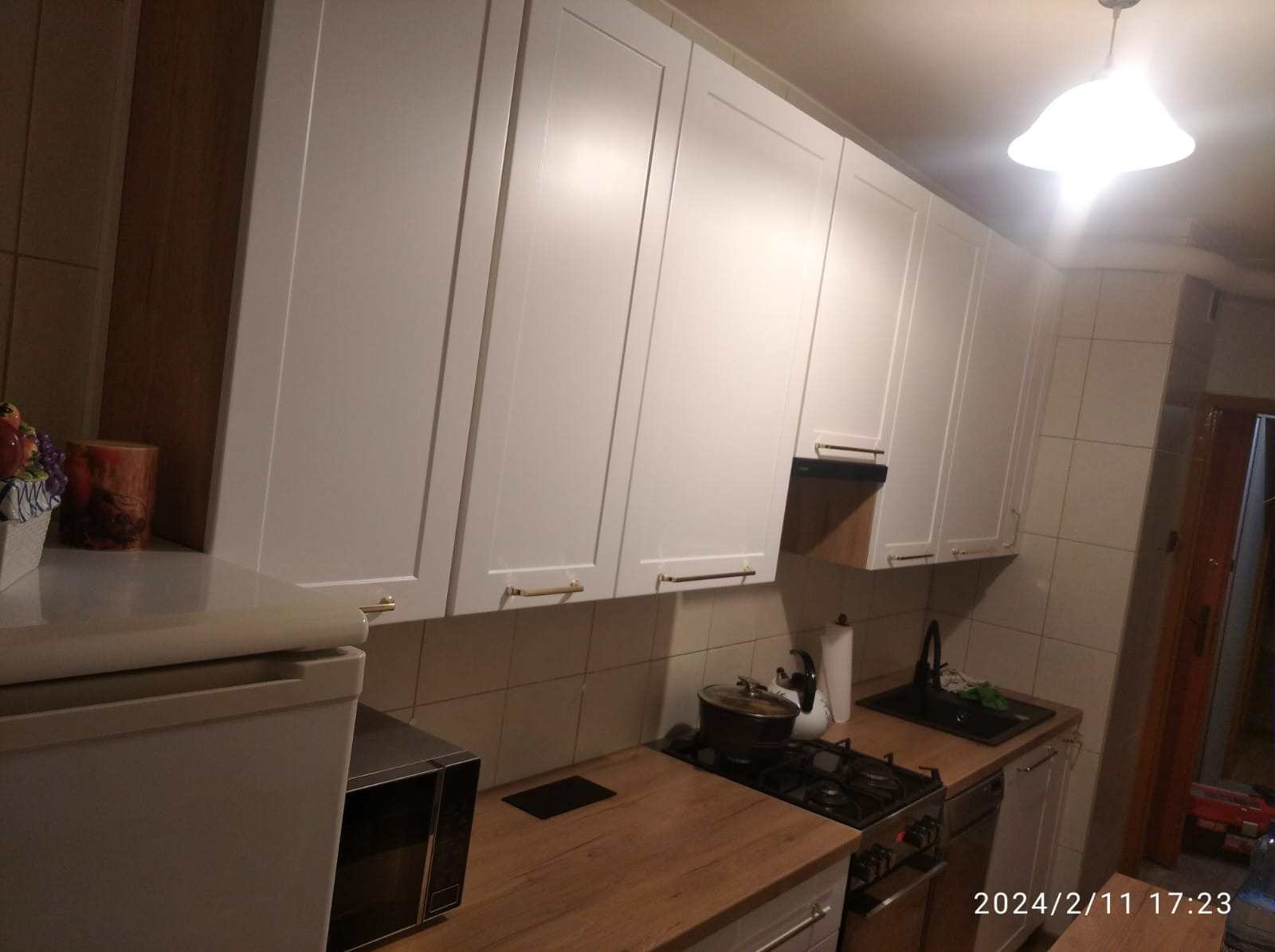 Kuchnia po remoncie w Gościno: białe szafki wiszące z poziomymi, złotymi uchwytami, drewniane blaty, płytki na ścianie i widoczny fragment kuchenki gazowej.