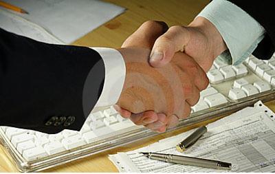 Uścisk dłoni dwóch osób nad klawiaturą komputera, dokumentami i długopisem. Elementy sugerują zawarcie umowy lub porozumienia biznesowego.