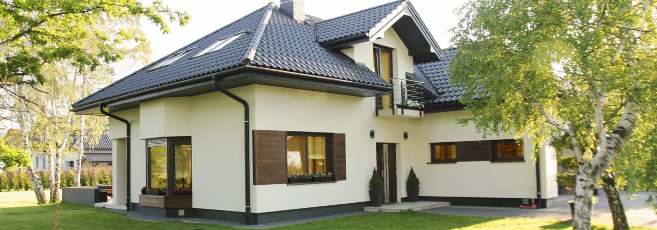 Elewacja domu jednorodzinnego z ciemną dachówką, drewnianymi okiennicami i balkonem, otoczona zielenią.