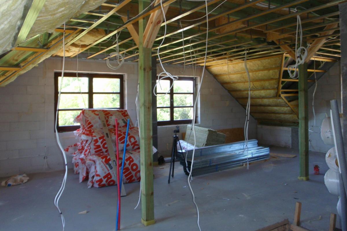 Poddasze w trakcie adaptacji: widoczne okna, izolacja z wełny mineralnej na skośnej ścianie, konstrukcja sufitu podwieszanego, materiały budowlane składowane na podłodze, oraz przewody elektryczne.