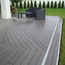 Szary taras z desek kompozytowych z meblami ogrodowymi z technorattanu, widok na trawnik i rząd tuj w tle.
