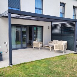 Egrocenter - Nowoczesne zadaszenie tarasu z ciemnoszarą metalową ramą i przezroczystym dachem, przylegające do jasnego domu. Pod nim jasne meble ogrodowe na płytkach, obok panel lamelowy.