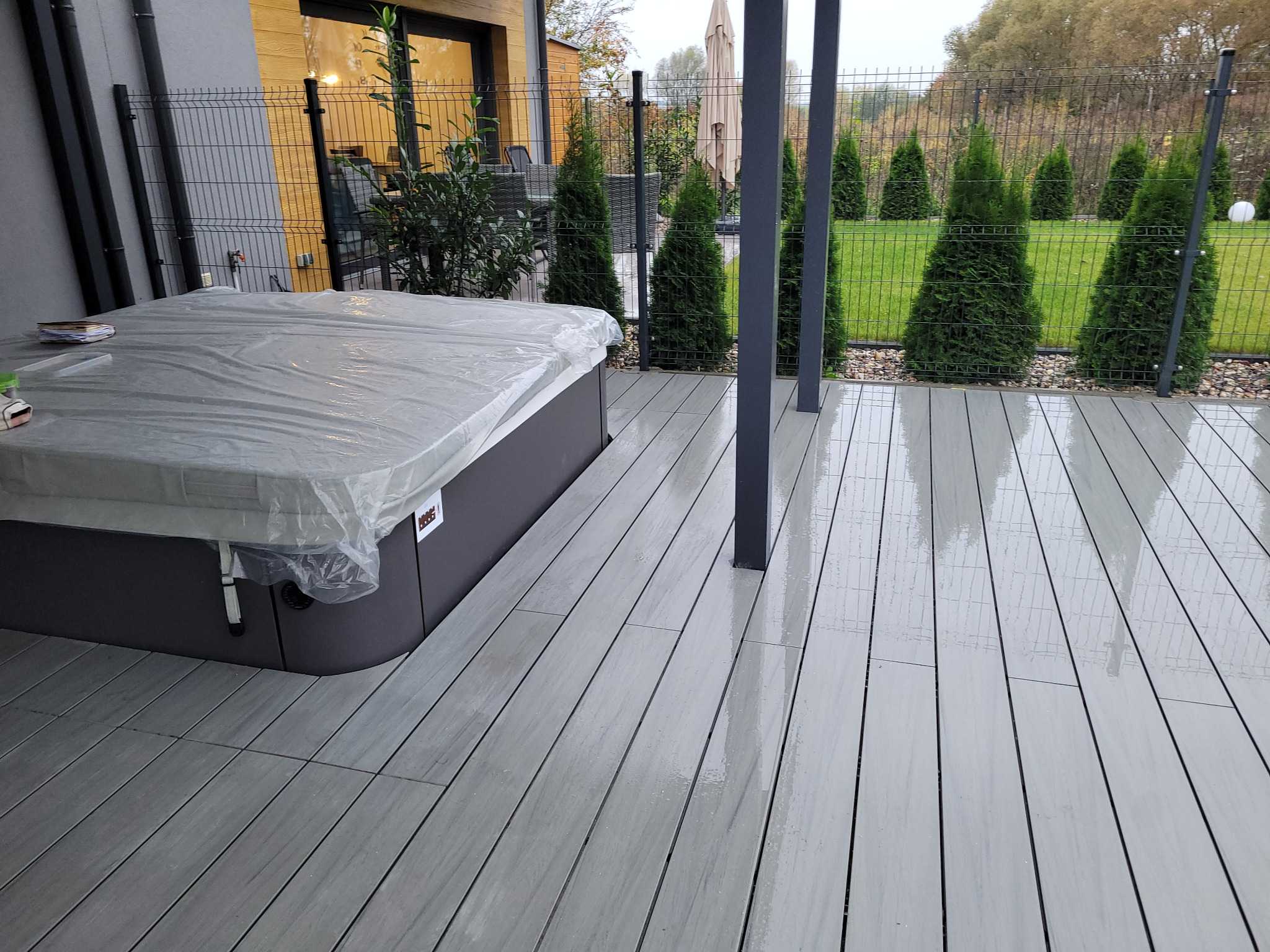 Szary taras z desek, przykryta folią wanna spa, w tle rząd tuj i metalowe ogrodzenie.
