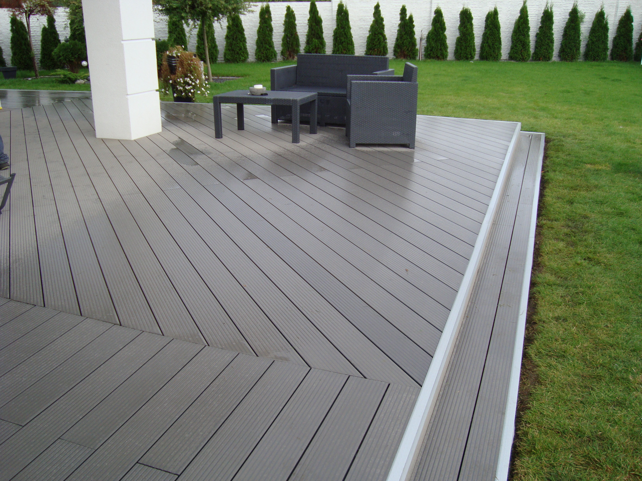Szary taras z desek kompozytowych z meblami ogrodowymi z technorattanu, widok na trawnik i rząd tuj w tle.