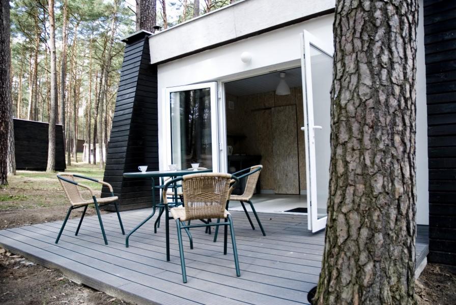 Nowoczesny domek letniskowy z tarasem kompozytowym i zielonymi krzesłami, otoczony lasem sosnowym, z widocznymi filiżankami na stole.