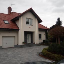 Bud-med - Elewacja domu jednorodzinnego z czerwoną dachówką, otynkowana na kremowo, z szarym brukowanym podjazdem i zadbanym ogrodem w pochmurny dzień.