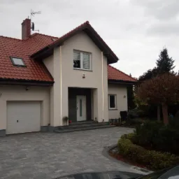 Elewacja domu jednorodzinnego z czerwoną dachówką, otynkowana na kremowo, z szarym brukowanym podjazdem i zadbanym ogrodem w pochmurny dzień.