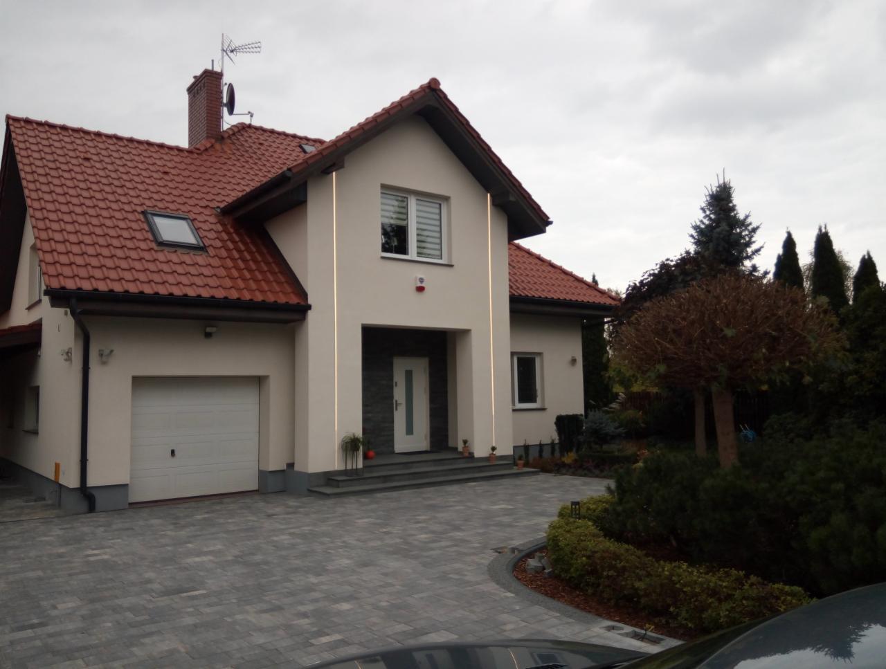 Elewacja domu jednorodzinnego z czerwoną dachówką, otynkowana na kremowo, z szarym brukowanym podjazdem i zadbanym ogrodem w pochmurny dzień.